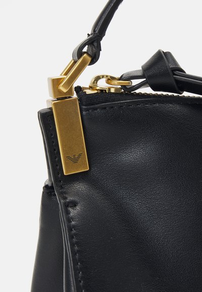Melnas ādas rokassoma ar teksturētu virsmu, zelta toņa aksesuāriem, rāvējslēdzēju un ar zīmolu akcenta detaļu netālu no siksnas.