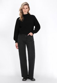 Sort strikket sweater med høj hals, parret med højtlivet, vide sorte jeans. Modellen står med den ene hånd i lommen.