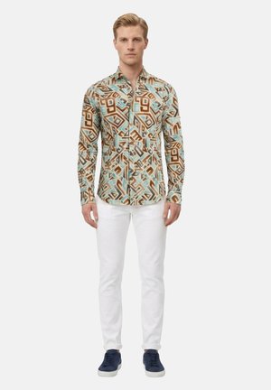 Uomo in piedi che indossa una camicia a maniche lunghe con motivo geometrico multicolore, pantaloni bianchi e scarpe da ginnastica blu navy su sfondo semplice.