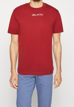 Homme portant un t-shirt rouge à manches courtes avec le texte « Marc O Polo » sur la poitrine et un pantalon bleu clair, debout devant un fond blanc uni.