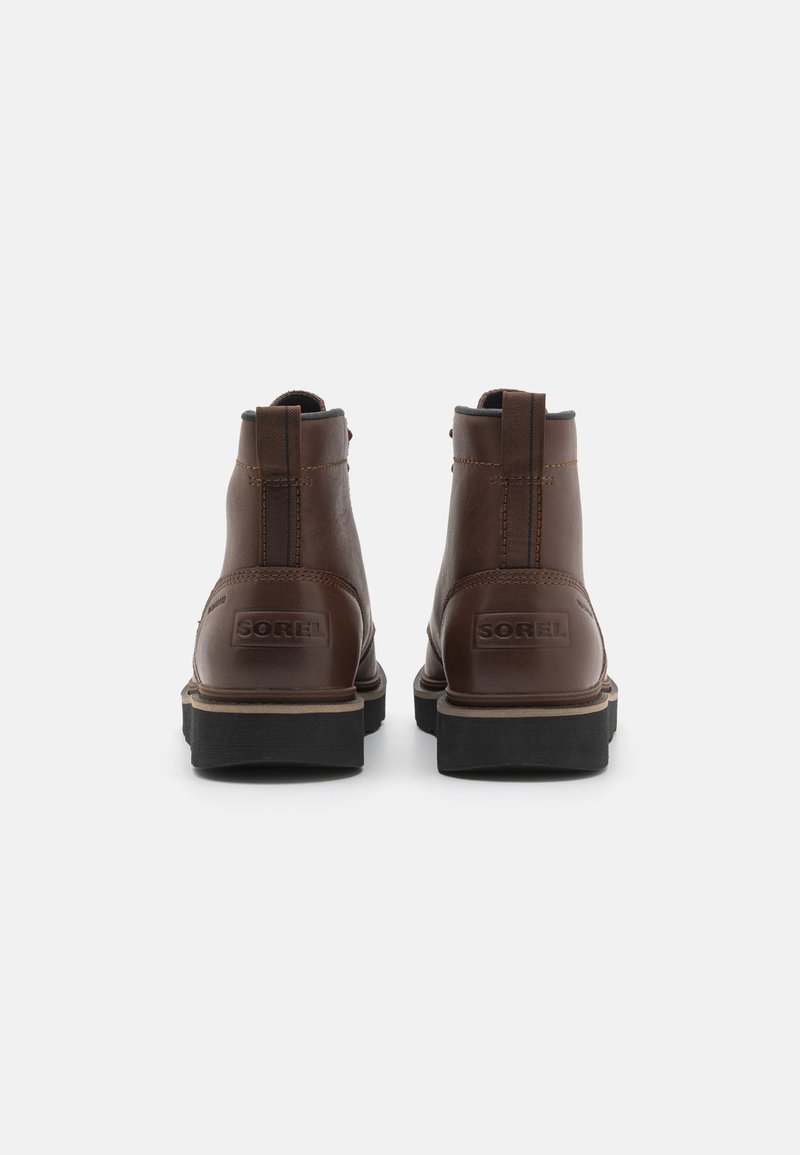 Bottines en cuir marron avec une finition lisse, des panneaux latéraux élastiques et une semelle en caoutchouc noir surélevée ; logo Sorel sur le talon.