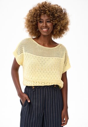 Kobieta o kręconych włosach, ubrana w żółty dzianinowy top i granatowe spodnie w paski z wysokim stanem, uśmiecha się z jedną ręką w kieszeni.