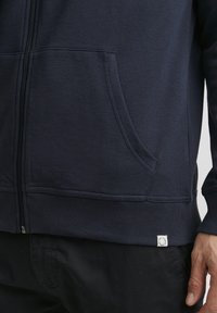 Sweat à capuche zippé bleu marine en matière douce, doté d'une poche avant, d'un ourlet côtelé et d'un petit patch logo en bas à gauche.