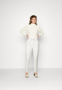 Witte geribde coltrui gecombineerd met witte slim-fit broek met een elastische taille en twee zijzakken. Neutrale hakken.