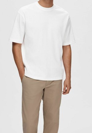 Man draagt een simpel wit T-shirt met korte mouwen en beige broek, met één hand in de zak, tegen een effen achtergrond.