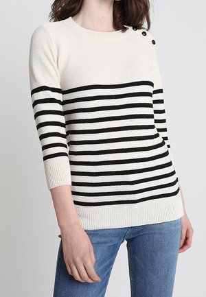 Femme portant un pull en tricot blanc avec des rayures horizontales noires et trois boutons noirs sur l'épaule gauche, assorti à un jean bleu.
