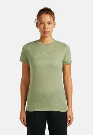 Jonge vrouw met krullend haar, gekleed in een lichtgroen T-shirt met een ontwerp van een witte bergketen, een halvemaan en sterren, staand tegen een witte achtergrond.