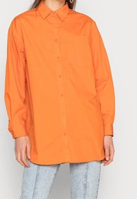 Donna che indossa una camicia arancione oversized con bottoni e taschino, abbinata a jeans azzurri, in piedi contro uno sfondo semplice.