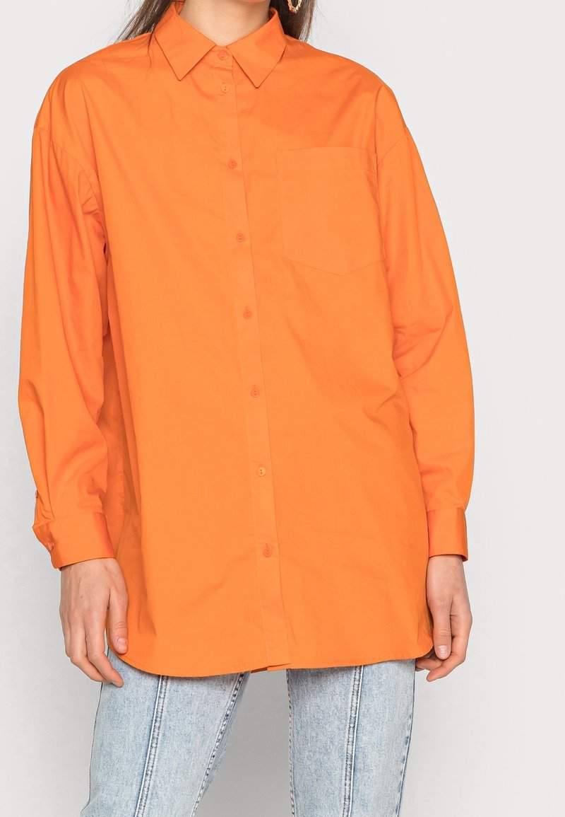 Donna che indossa una camicia arancione oversized con bottoni e taschino, abbinata a jeans azzurri, in piedi contro uno sfondo semplice.
