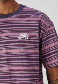 Svītrainā kokvilnas T-kreklā purpura toņos ir apaļa kakla izgriezuma forma un neliels balts Nike SB logo kreisajā krūtī.
