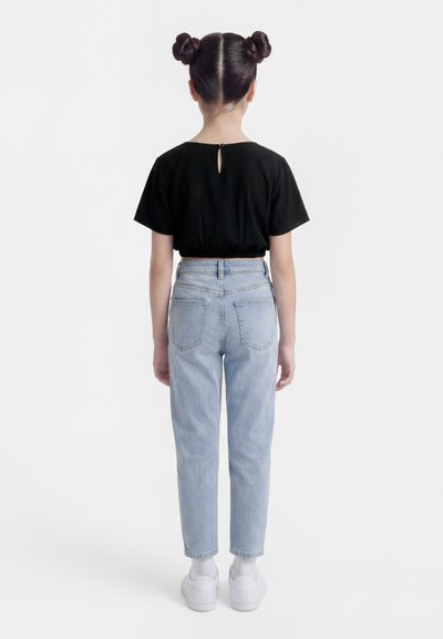 Παιδί με δύο κότσους στα μαλλιά στέκεται με την πλάτη προς τα εμπρός, φορώντας μαύρο κοντό μπλουζάκι, ανοιχτό μπλε τζιν, λευκές κάλτσες και λευκά αθλητικά παπούτσια.