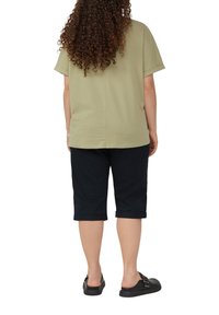 T-shirt verde oliva a maniche corte con taglio dritto; abbinato a pantaloni corti neri fino al ginocchio. Modello che indossa scarpe slip-on nere.