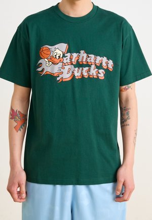 T-shirt vert avec un canard de dessin animé orange et blanc tenant un ballon de basket et le texte "Carhartt Ducks" porté par une personne avec des bras tatoués.