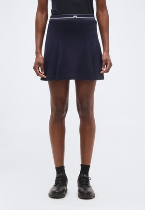 Person iført en navy plisseret nederdel med hvidt taljebånd, sort top, sorte sokker og sorte snøresko, stående mod en ensfarvet baggrund.