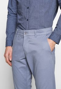 Man med ljusblå byxor och en blå mönstrad skjorta med knappar, med ena handen i fickan och den andra avslappnad vid sidan.