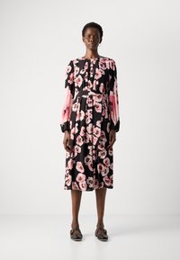 Mujer vestida con un vestido midi negro con estampado floral rosa, mangas largas y cinturón en la cintura, de pie con zapatos negros de hebilla sobre fondo blanco.