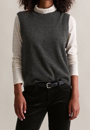 Person trägt einen dunkelgrauen ärmellosen Pullover über einem weißen karierten Rollkragenhemd und schwarzen Hosen mit schwarzem Gürtel.