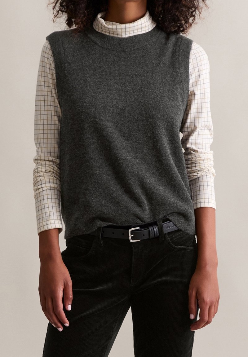 Personne portant un pull sans manches gris foncé sur une chemise à col roulé blanche à carreaux et un pantalon noir avec une ceinture noire.
