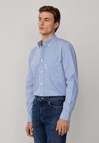 Hombre con una camisa de botones de rayas verticales azul y blanca y pantalones vaqueros azul oscuro, de pie contra un fondo gris liso.