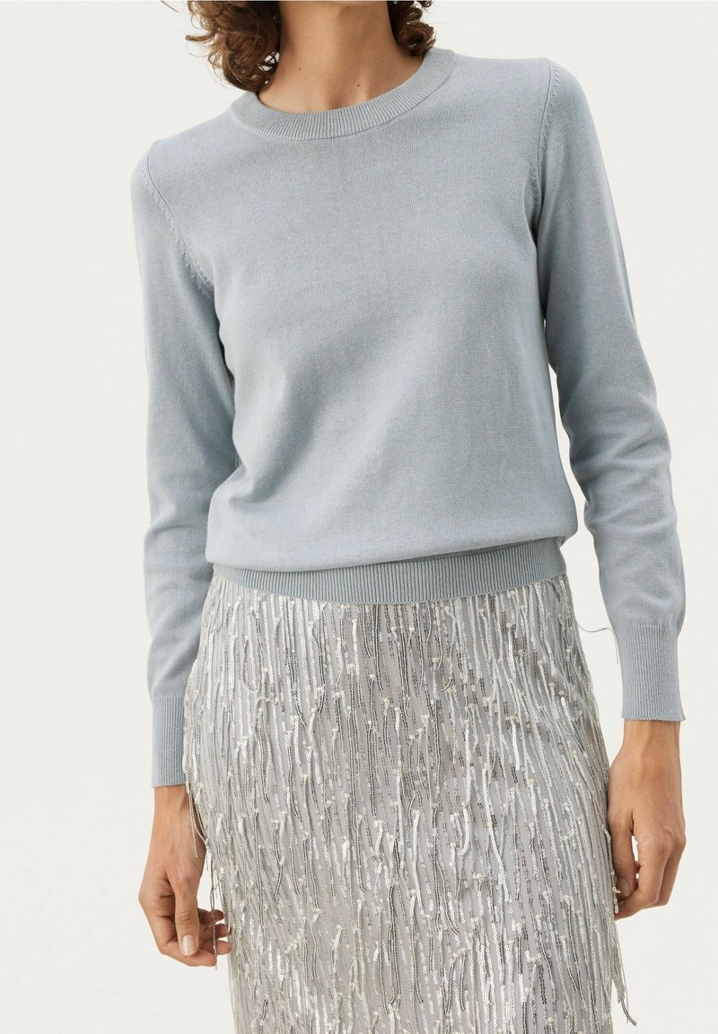 Maglione lavorato a maglia azzurro chiaro con maniche lunghe e scollo a girocollo, abbinato a una gonna argentata con frange e dettagli lucenti.