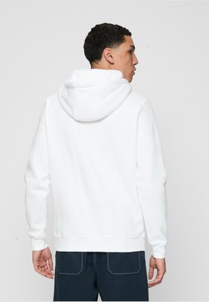 Muž v obyčejné bílé mikině s kapucí a tmavě modrých kalhotách, pohled zezadu na jednobarevném světlém pozadí.