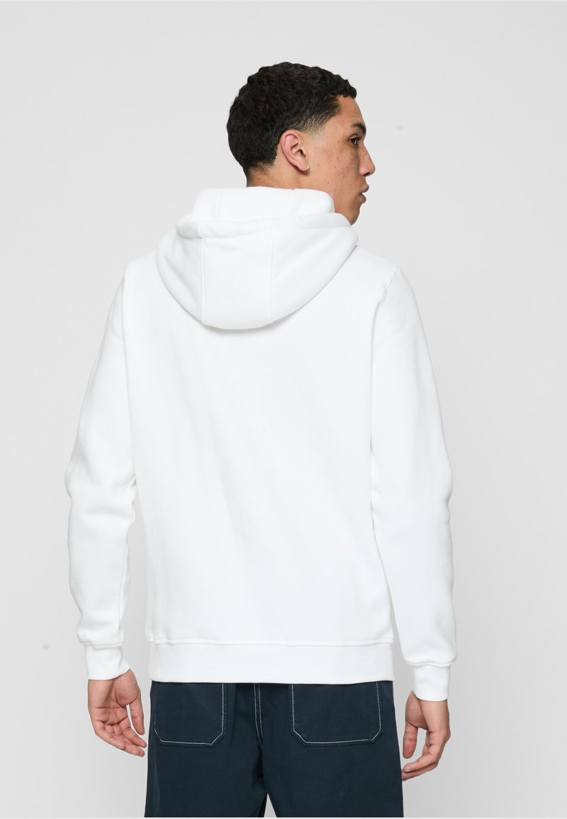 Uomo visto da dietro, indossa una felpa con cappuccio bianca semplice e pantaloni blu scuro, su uno sfondo chiaro e uniforme.