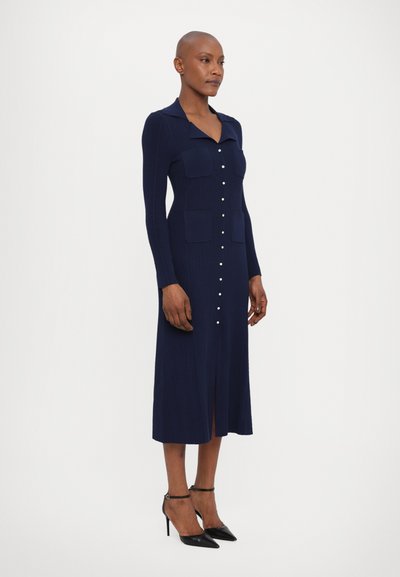 Abito a maglia a coste blu navy con maniche lunghe, chiusura frontale con bottoni, due tasche sul petto e orlo con spacco. Abbinato a tacchi neri e cinturini alla caviglia.