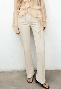 Beige flared trousers with a smooth texture, high waist, and side seams. Paired with brown sandals, set against a plain white background.