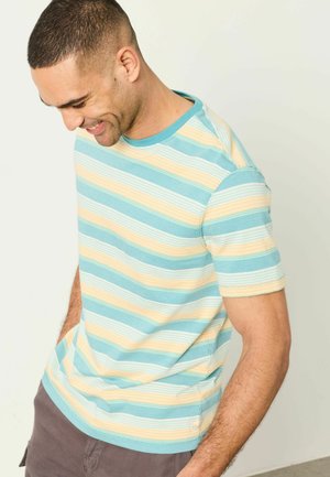 Man smiling and looking down wearing a short-sleeve, blue, yellow, and white striped shirt with hands in pockets against a plain background.