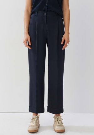 Navy high-waisted trousers with front pleats, foldable cuffs, and a button closure. Paired with beige sneakers for a neutral contrast.