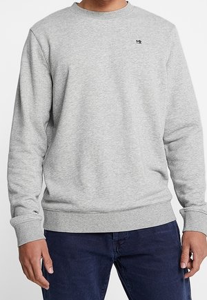 Mężczyzna ubrany w jasnoszary sweatshirt z okrągłym dekoltem z małym, czarnym haftowanym logo na piersi oraz ciemnoniebieskie dżinsy, stojący z luźno opuszczonymi rękami.