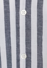 Bílá látka s tmavými vertikálními pruhy, na které jsou sešity dva bílé knoflíky ve vertikální řadě.