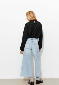 Haut noir à manches longues avec un détail de col volanté, associé à un jean larges bleu clair et des chaussures noires, sur un fond neutre.