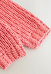 Spoži rozā adīta džempera piedurkne ar ribotu aproci un fakturētu valdziņu rakstu, izceļot mīkstu, biezu dziju.