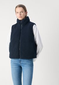 Gilet in pile blu navy con colletto alto, chiusura con zip e tasche laterali. Texture morbida, design senza maniche, indossato sopra una maglietta bianca.