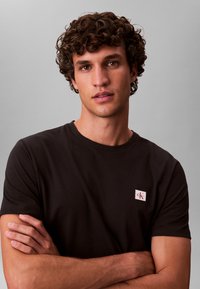 Must puuvillane crew-neck T-särk, millel on vasakul rinnal väike valge Calvin Kleini logo silt. Modellil on lokkis juuksed ja ta on käed risti.