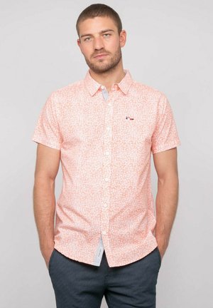 Homme aux cheveux courts et barbe portant une chemise rose à manches courtes à motifs floraux et un pantalon foncé à motifs, mains dans les poches.