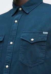 Blauw button-up shirt met parelachtige drukknoopjes, borstzakje aan de rechterkant en een klein genaaid JACK & JONES label.