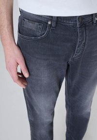 Jean en denim gris foncé avec une coupe slim, comprenant un design à cinq poches, des boutons en métal et un décoloration subtile au niveau des cuisses.