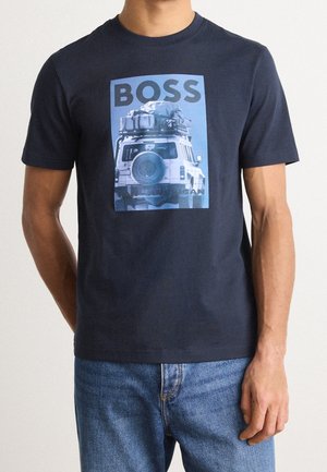 Man wearing navy blue T-shirt with graphic of off-road vehicle carrying luggage and text "BOSS" on front, paired with blue jeans.