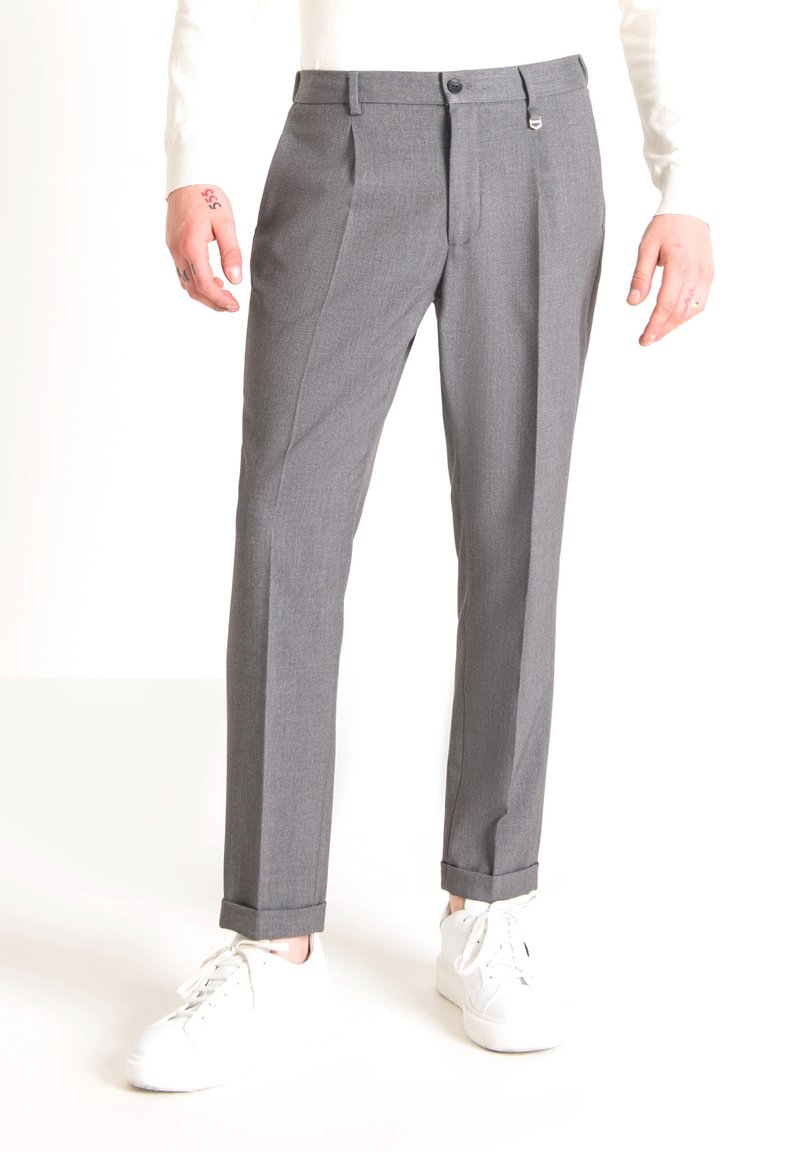 Personne portant un pantalon gris sur mesure avec revers, associé à des baskets blanches et un haut blanc à manches longues, debout devant un fond blanc.