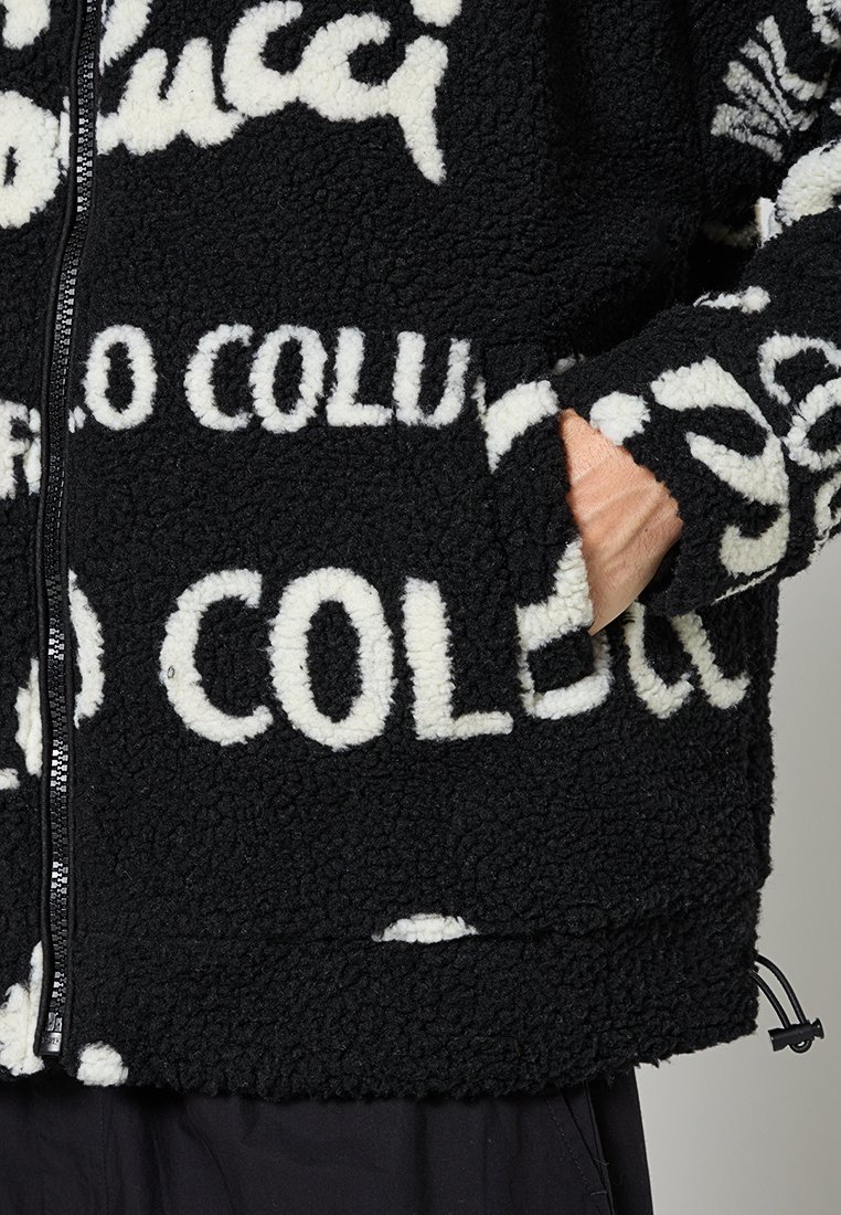 Schwarze Fleecejacke mit weißem Textdesign. Verfügt über einen Reißverschluss vorne, Seitentaschen und einen strukturierten, groben Stoff.