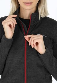 Giacca nera con zip e finitura testurizzata, caratterizzata da una zip rossa e un logo sul lato sinistro. Le mani afferrano il tirante della zip.