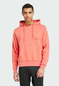 Koraalkleurige hoodie met een voorzak en geribbelde boorden. Bevat de subtiele opdruk "Adidas Football" op de borst. Soepele stoftextuur.