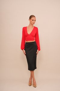 Rood lang mouw cropped top met een diepe V-hals, gecombineerd met een zwarte getailleerde midi rok met een split aan de voorkant. Het model draagt gouden hakken.