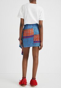 Nainen seisoo selkä cameras kohti, yllään valkoinen t-paita, sinivalkoraidallinen hame oransseilla kuoseilla ja punaiset fringepohjalliset sandaalit.