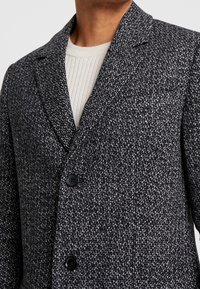 Blazer texturizado en gris y negro con solapa inglesa, dos botones frontales y un sutil patrón tejido. Llevado sobre un suéter ligero de canalé.