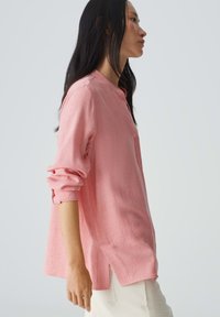 Camicetta lunga a maniche con texture di colore rosa chiaro, dotata di colletto e fessure laterali, caratterizzata da una vestibilità rilassata e maniche arrotolate.