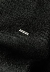 Tela de punto negra con un patrón texturizado, que cuenta con una pequeña etiqueta metálica con la palabra "SUPERDRY" sujeta de forma segura.