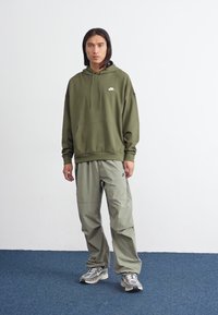 Ένα άτομο με μακριά μαλλιά στέκεται σε χαλαρή στάση, φορώντας μια ελιά πράσινη φούτερ, αντίστοιχες παντελόνες και αθλητικά παπούτσια, μπροστά σε έναν απλό φόντο.